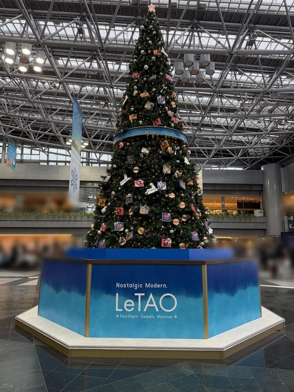 新千歳空港のセンタープラザに立つ大きなクリスマスツリー。お菓子の箱の形の飾りがたくさん付き、青い台にLeTAO Nostalgic Modern.の文字