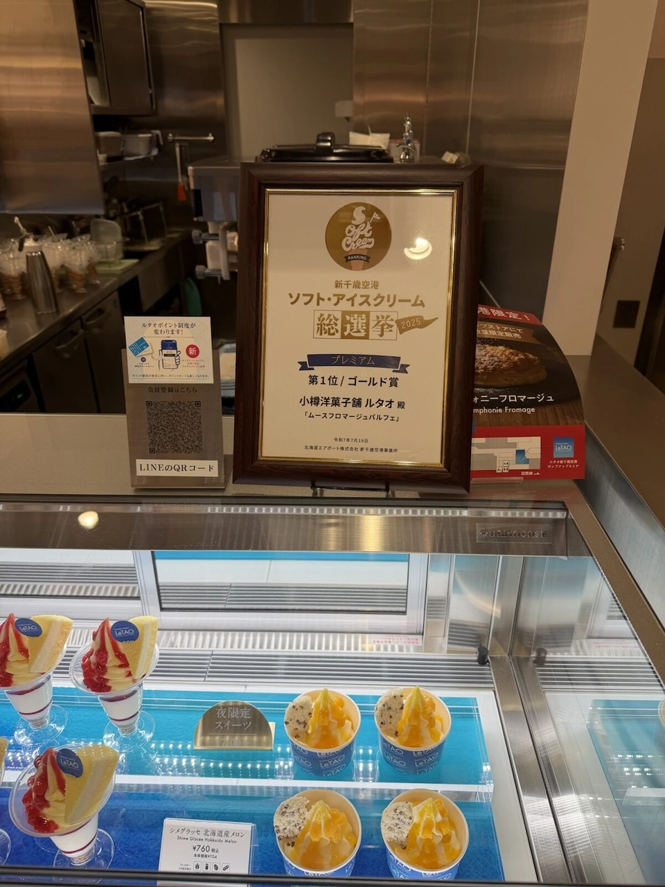 新千歳空港のルタオの店頭。ソフト・アイスクリーム総選挙2025プレミアム部門第1位ゴールド賞の額と、ショーケースに並ぶパルフェやメロンのグラッセ