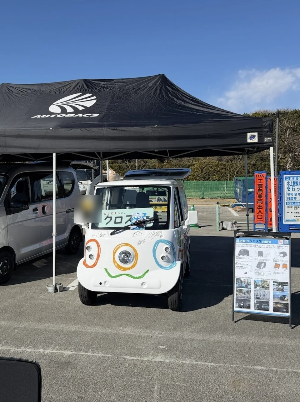 オートバックスの黒いテントの下に置かれた小さな白いEV。前面に顔のような絵とサイン、横に超小型EVクロスケの紹介の看板