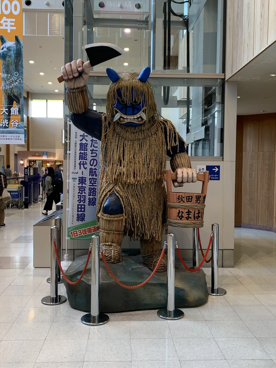 大館能代空港の到着ロビーに立つ大きななまはげの像。片手に包丁を掲げ、秋田男鹿なまはげと書かれた木の桶を持つ。横に大館能代と東京羽田線の幕