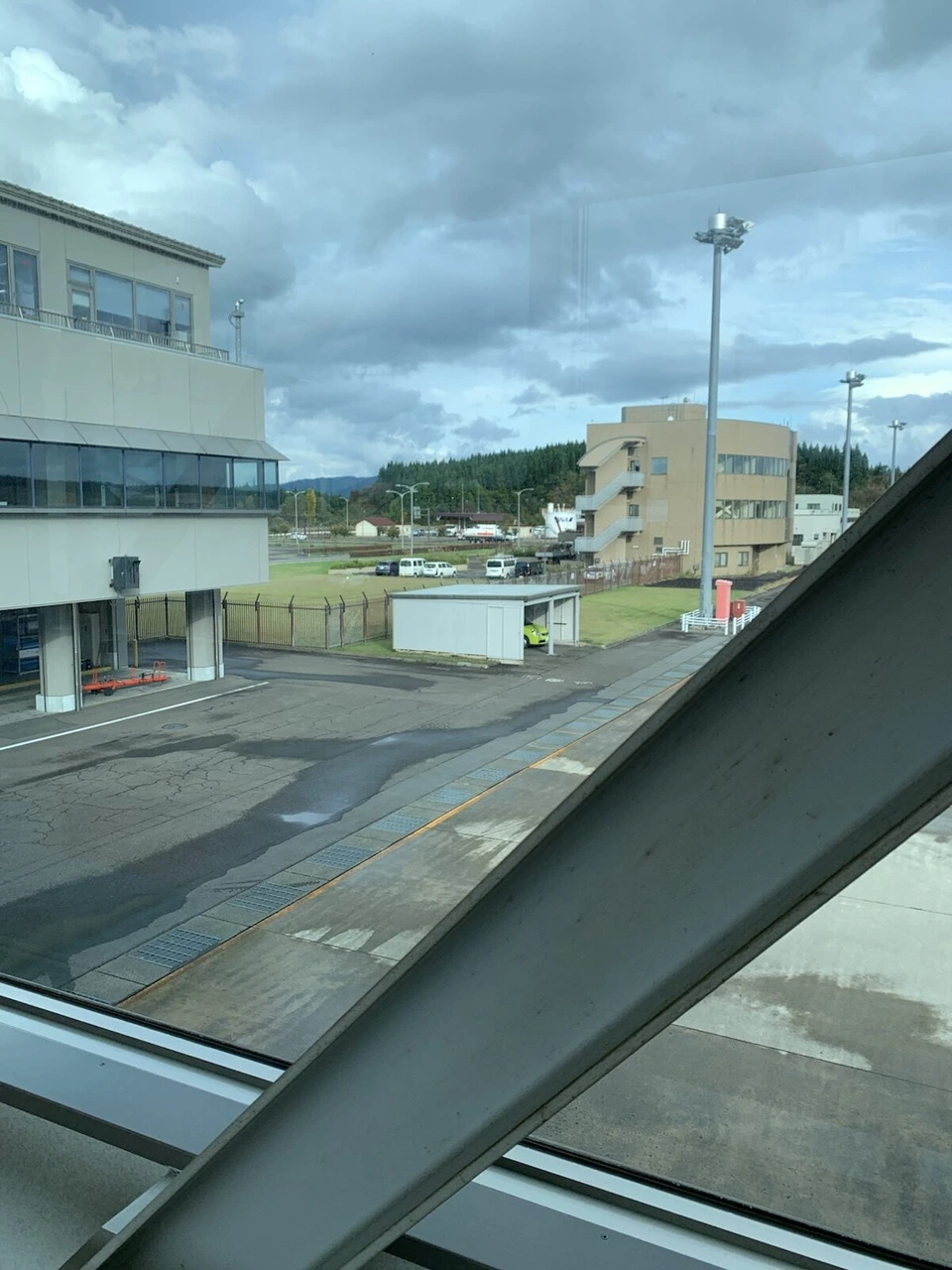 搭乗待合室の窓から見た空港の敷地。管理棟のような建物と照明塔、その奥に杉林。手前に窓枠の斜めの柱