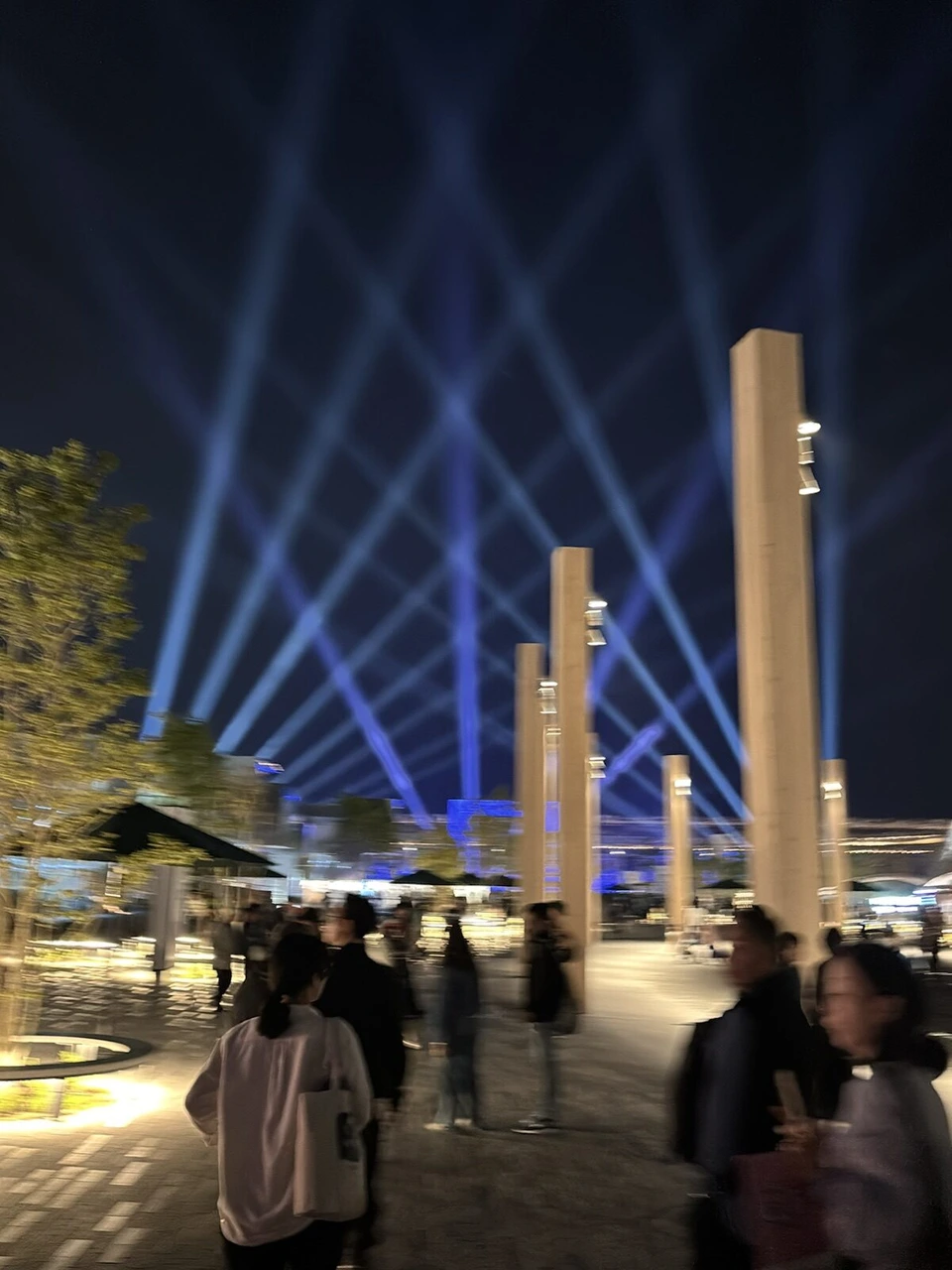 大阪・関西万博の夜空に交差する青いサーチライトの光と、ライトアップされた木の柱