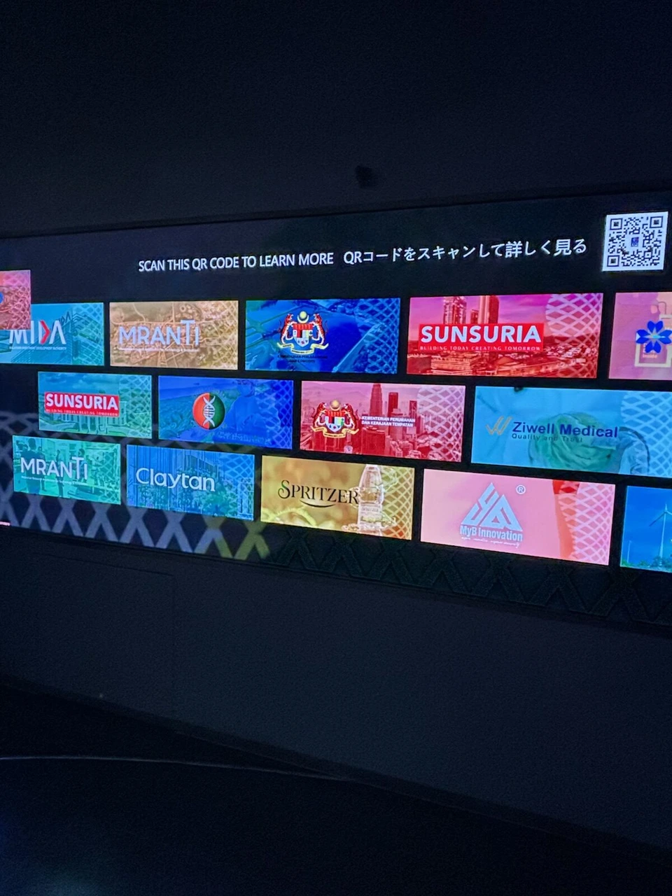 大阪・関西万博マレーシア館の暗い展示室の壁に並ぶ、マレーシアを紹介するカラフルな画面