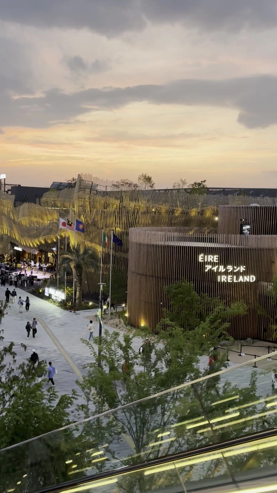 夕焼けの空の下の大阪・関西万博アイルランド館。木の縦格子の丸い建物とÉIRE アイルランド IRELANDの文字、手前にエスカレーターの手すり
