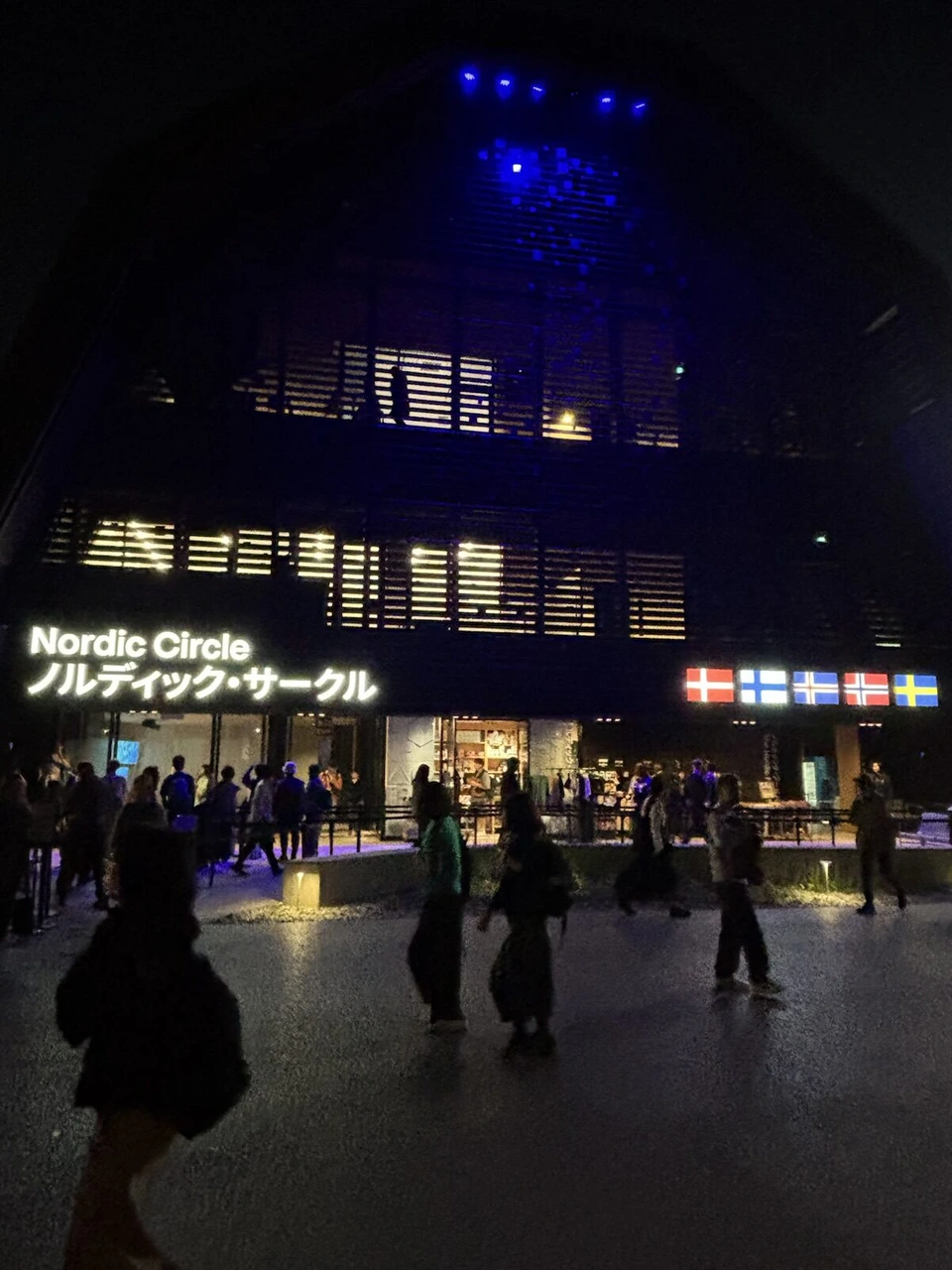 夜のノルディック・サークル館。青く光る外壁と、北欧5か国の国旗、入口に集まる人