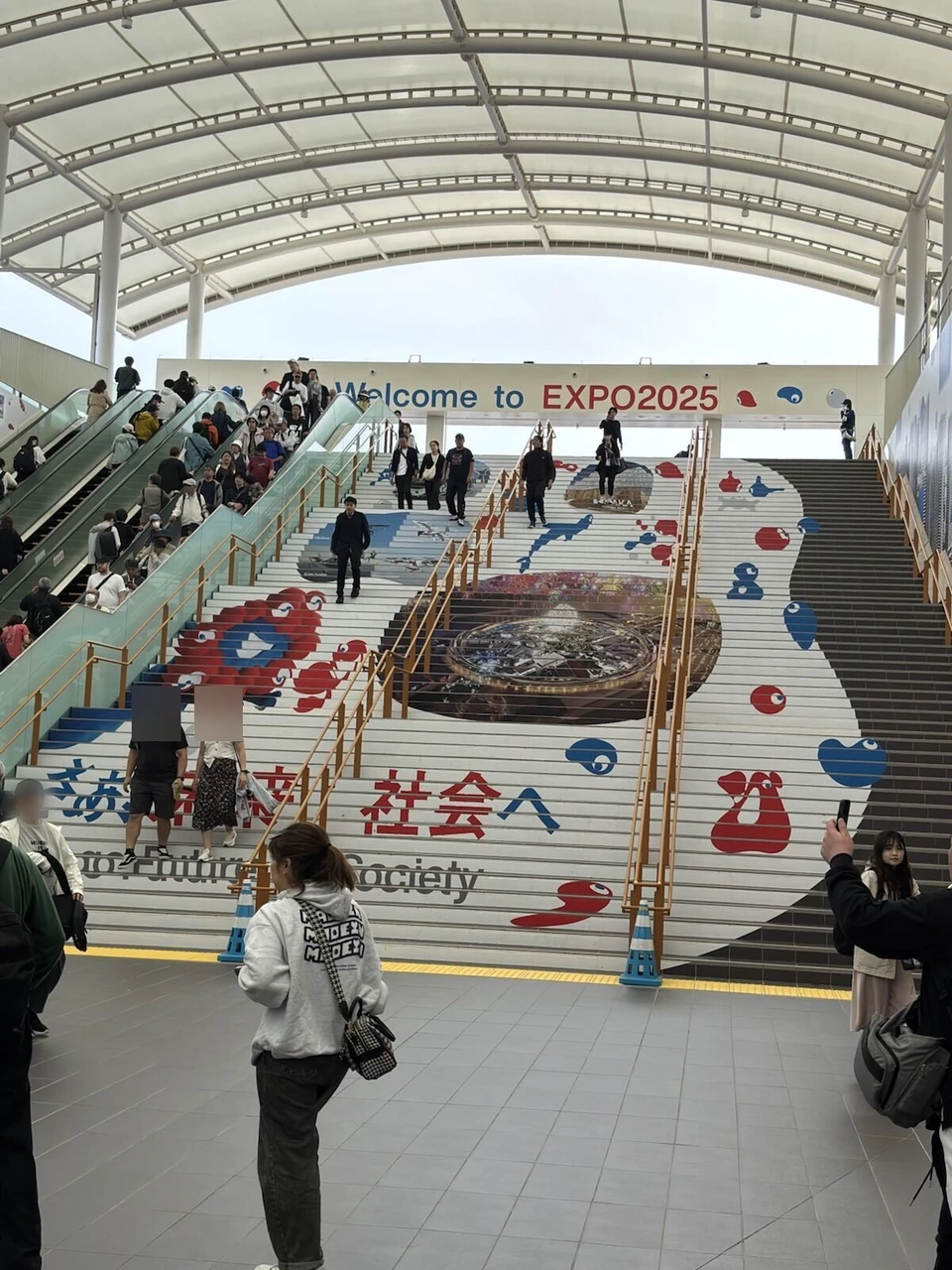 大阪・関西万博の東ゲート前の屋根付きの大きな階段。段に万博のロゴと絵、上にWelcome to EXPO2025の横断幕