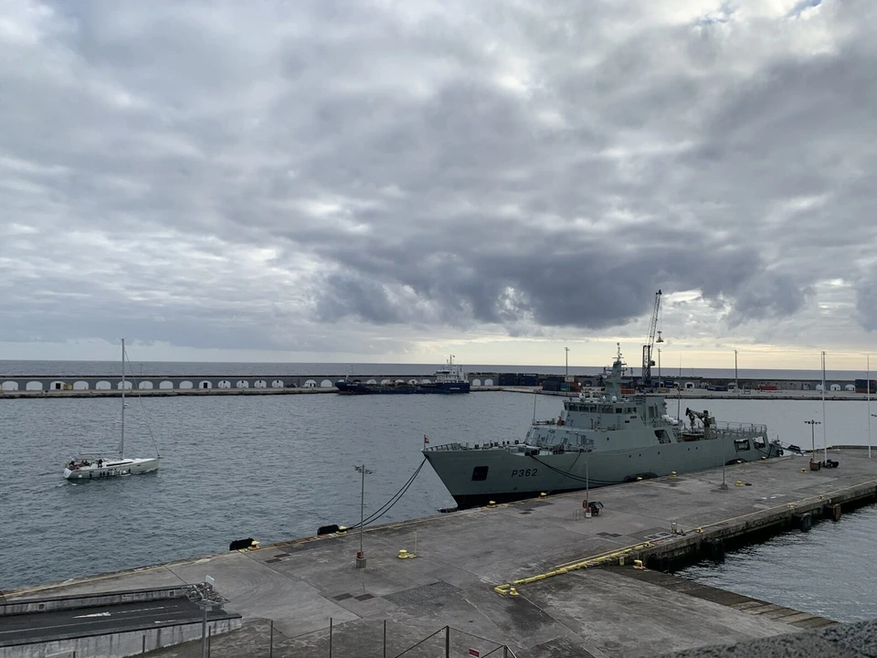 港の岸壁に泊まった灰色の軍艦と、奥の長い防波堤、海に浮かぶ1隻のヨット