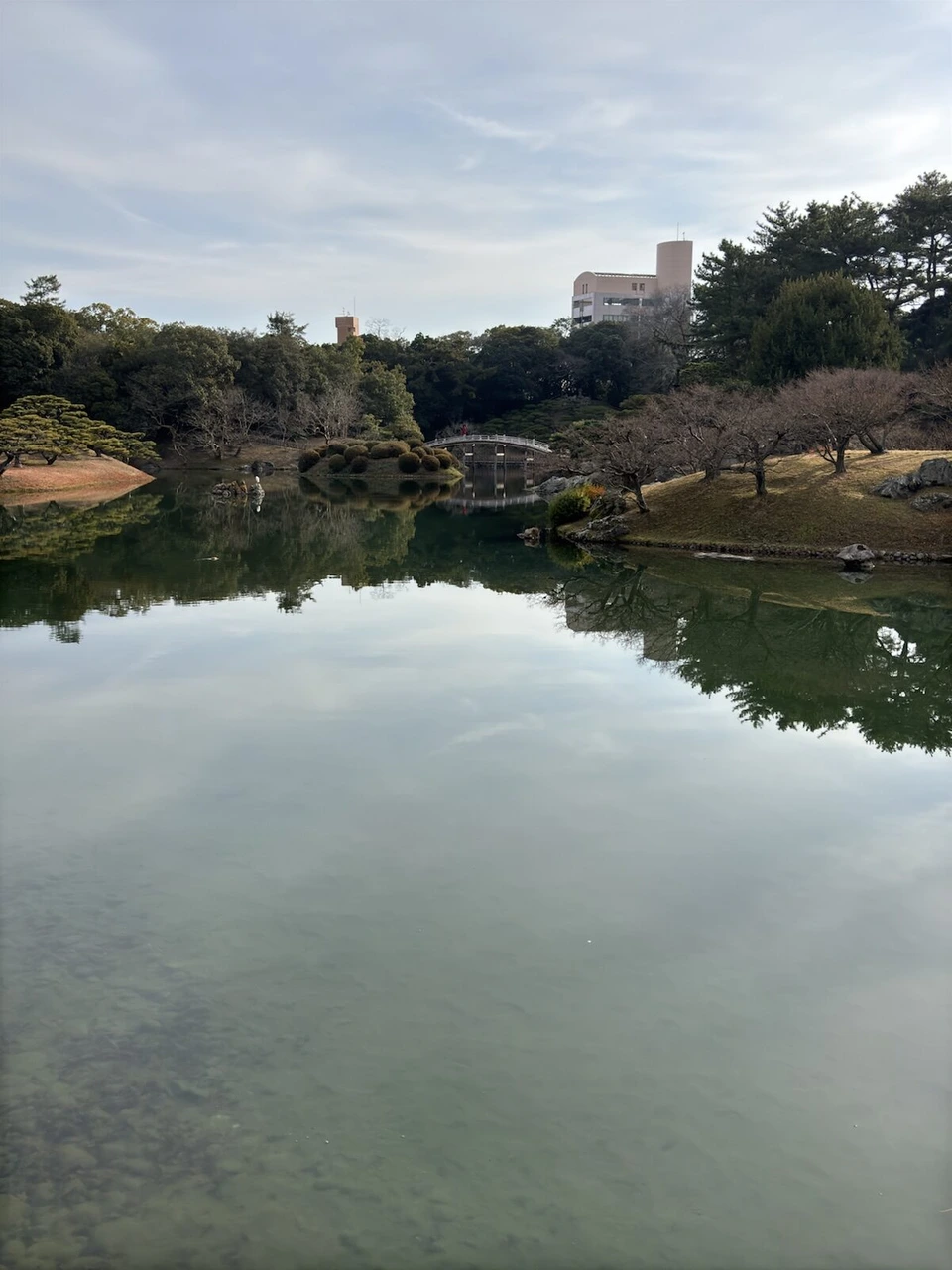 曇り空を映す栗林公園の南湖。対岸に松の木と小さく偃月橋、奥にビル