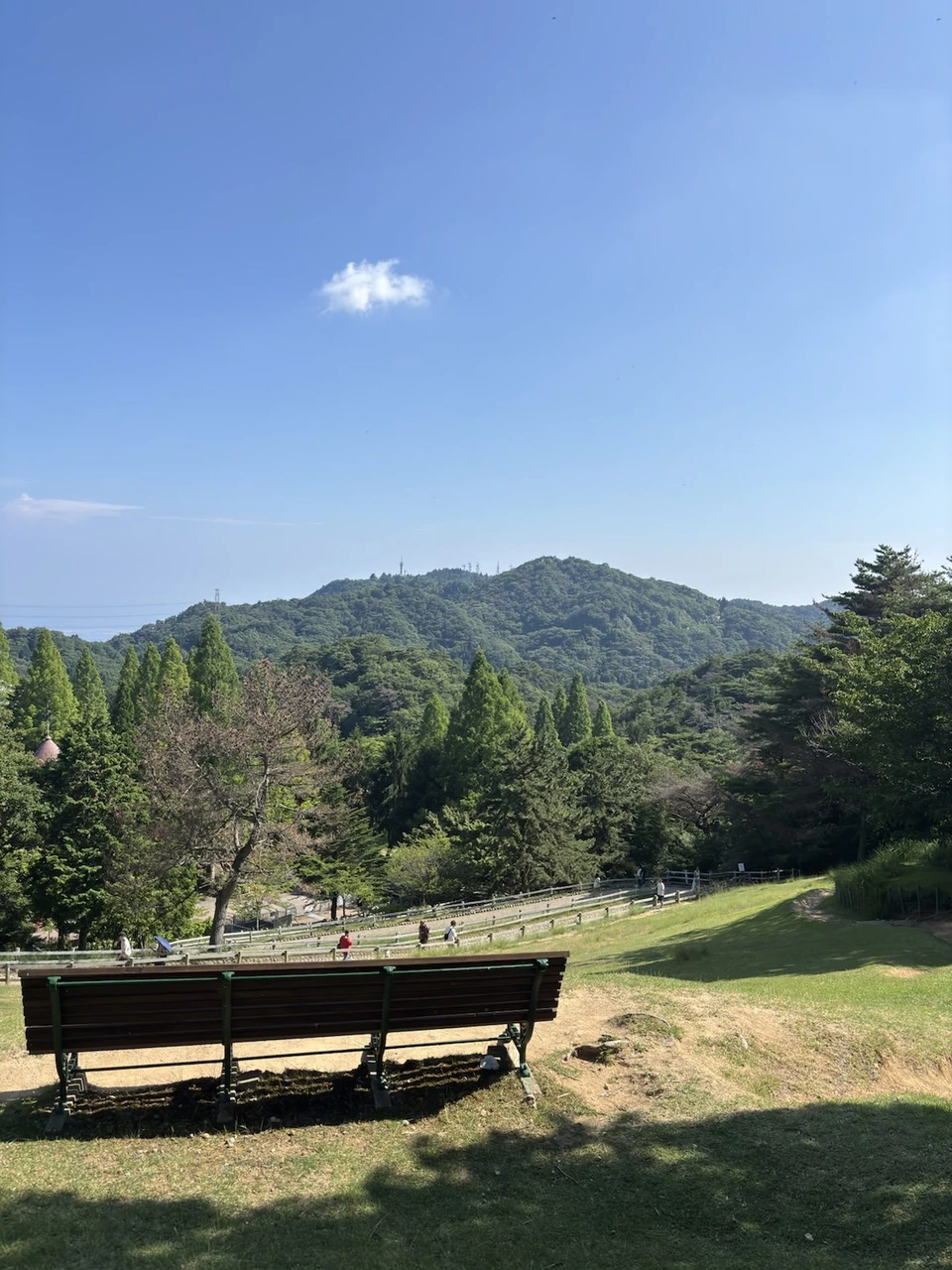 六甲山牧場の丘の上のベンチから見た景色。青空の下に緑の山が連なり、手前の芝生の斜面に柵と遊歩道