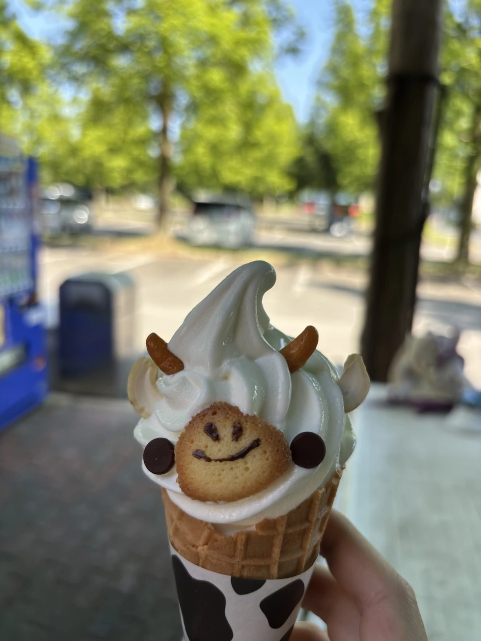 牛の顔に飾ったソフトクリーム。白いソフトに焼き菓子の顔、チョコの目と鼻、ナッツの角と耳。コーンの紙は白黒の牛柄。背景は木漏れ日の並木と駐車場