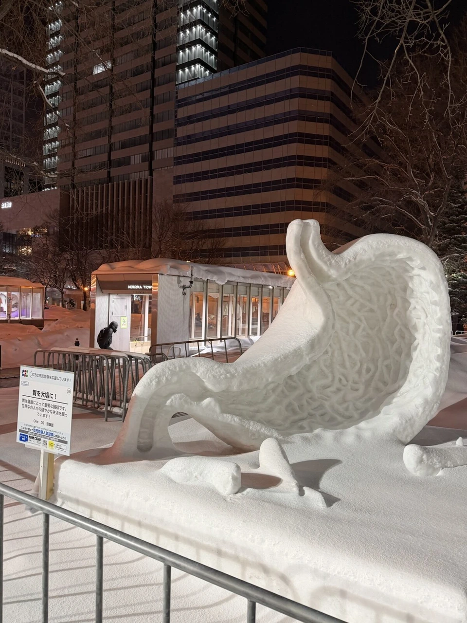 雪まつり大通会場の、大きく曲がった形の市民雪像と説明札、奥にビル