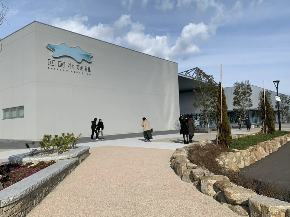 白い壁に四国水族館のロゴ。手前に歩道と石垣の植え込み