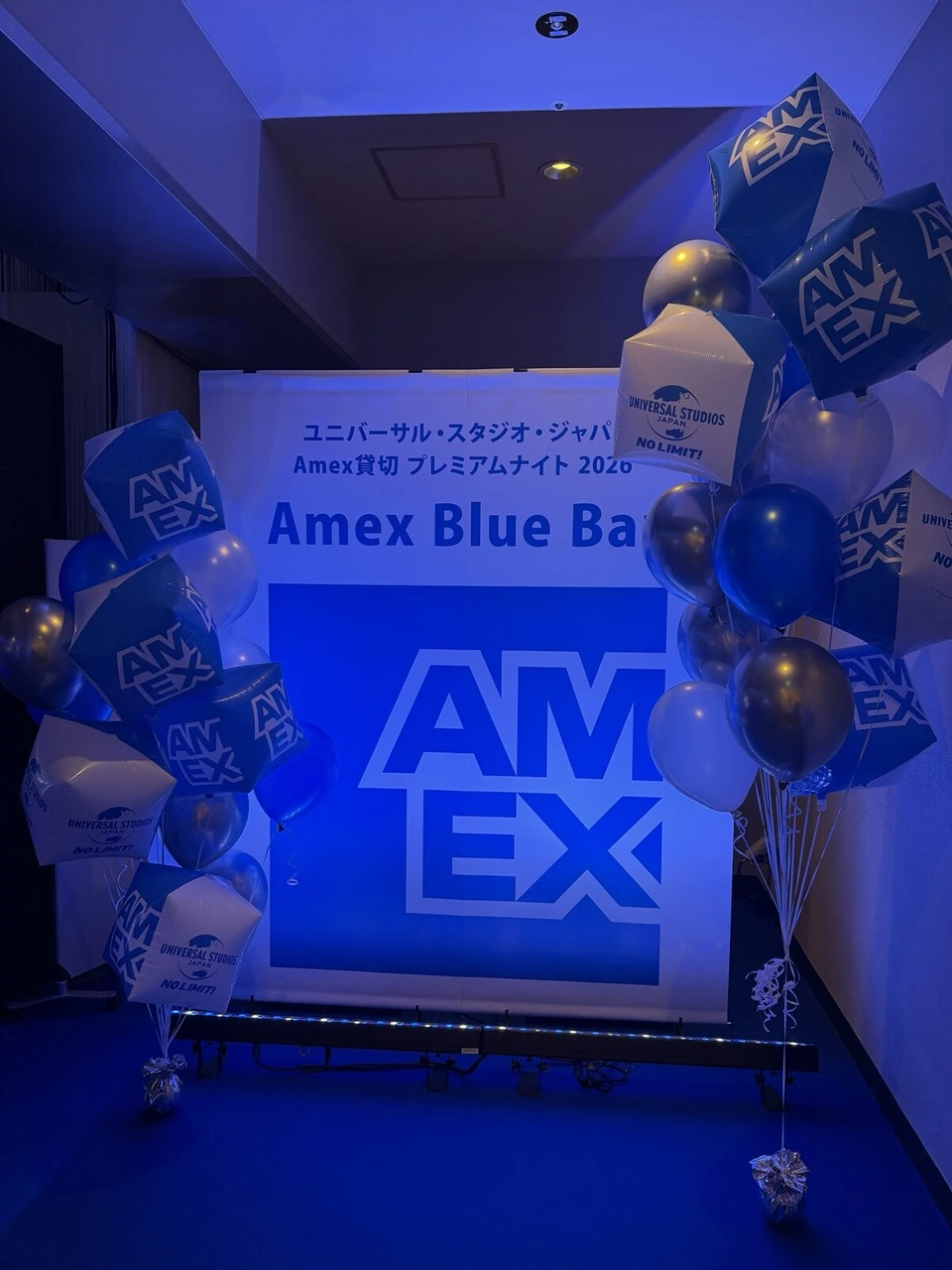 青い照明の中のAmex Blue Barの看板。AMEXのロゴ入りの風船がたくさん飾られている
