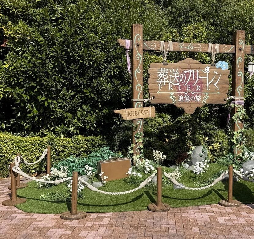 木の看板に「葬送のフリーレン 追憶の旅」と書かれたフォトスポット。手前に芝生とロープの柵