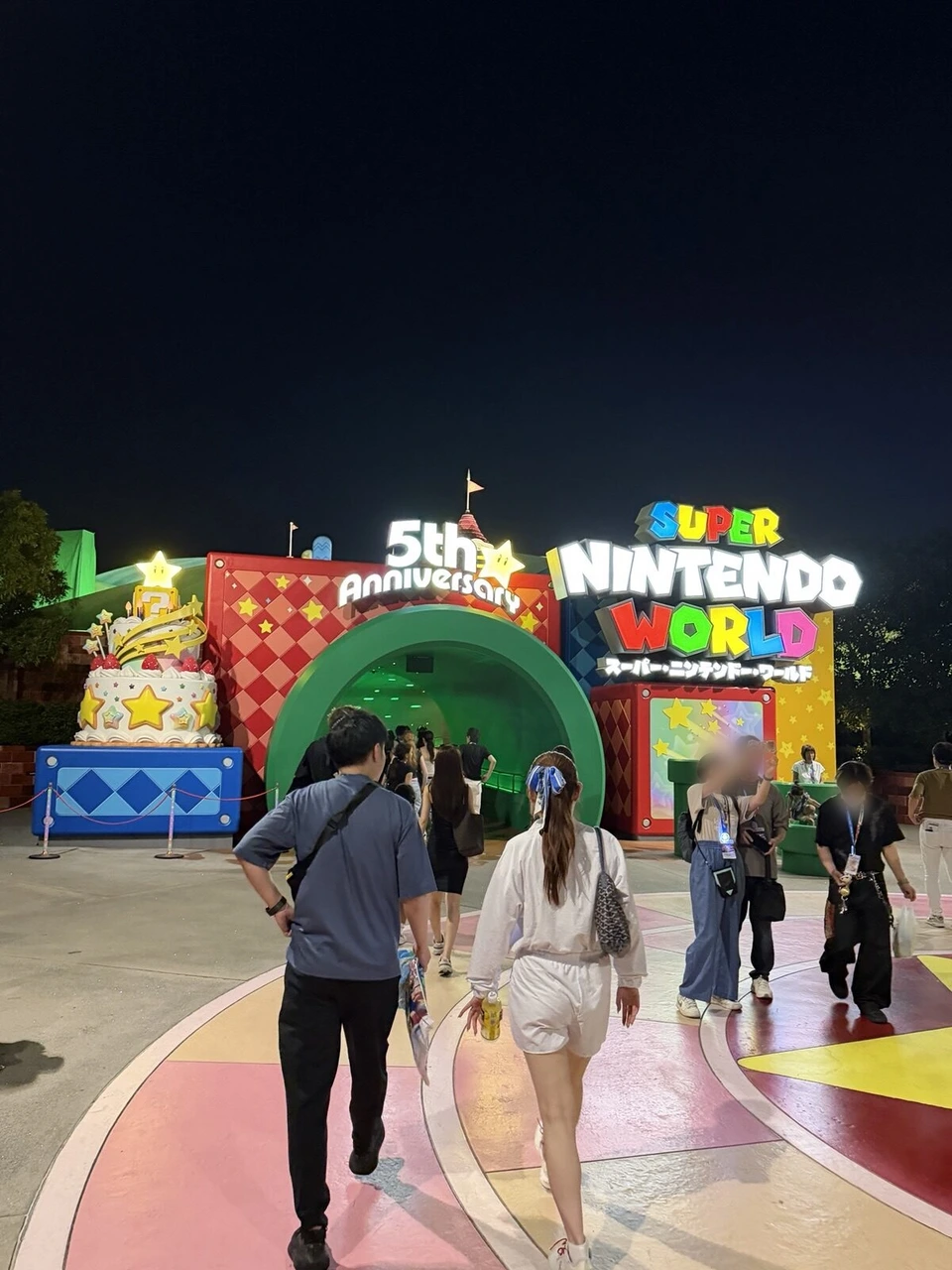 夜のスーパー・ニンテンドー・ワールドの入口。緑の土管のトンネルと5th Anniversaryの看板、ケーキの飾り