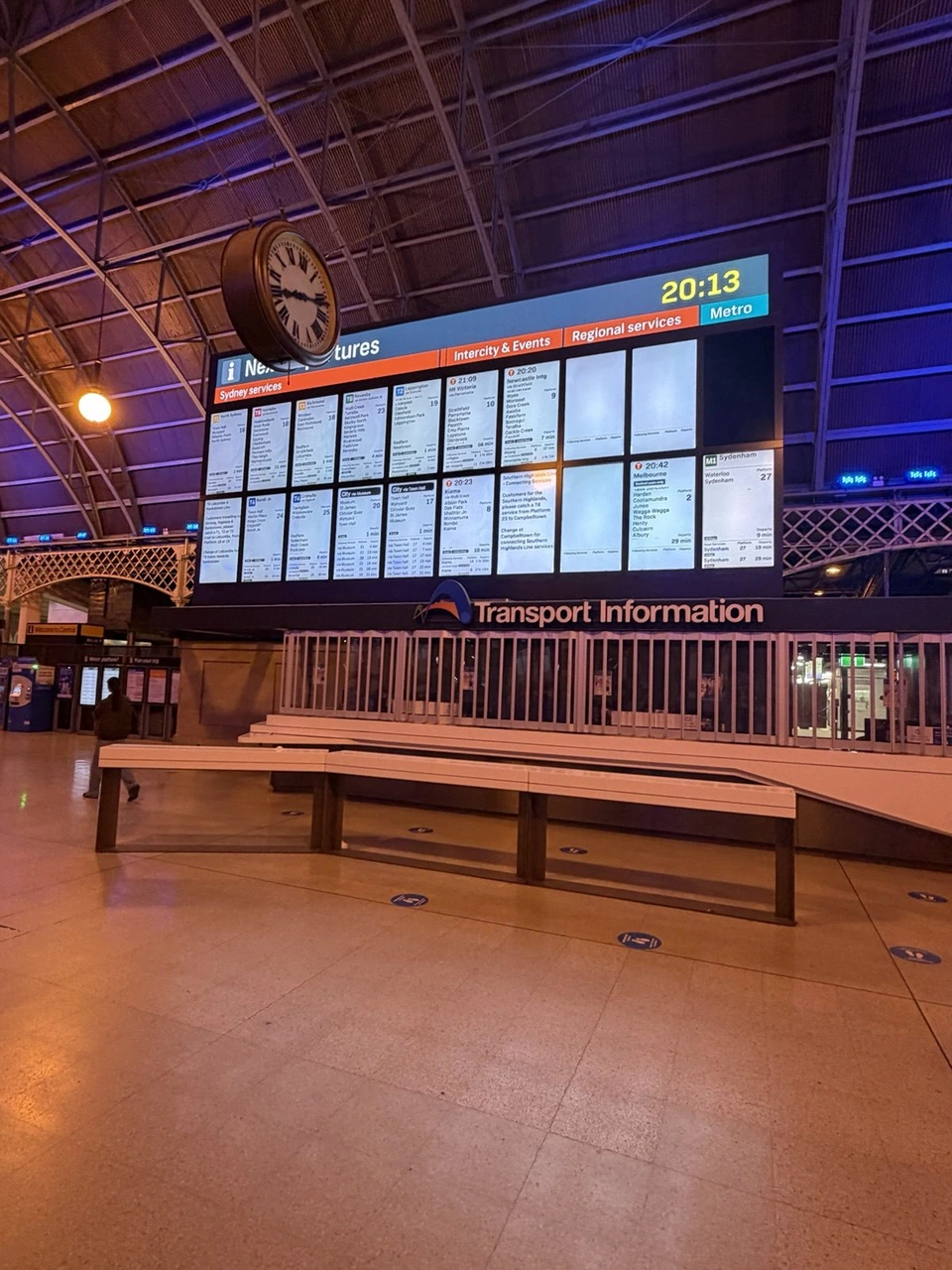 夜のシドニー中央駅のコンコース。アーチ形の屋根の下に大きな発車案内の画面と時計、手前に木のベンチ