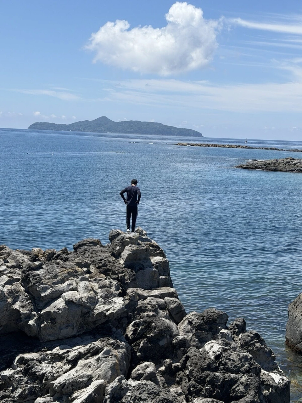 ごつごつした岩の先端に立つ人の後ろ姿と、沖の大きな島