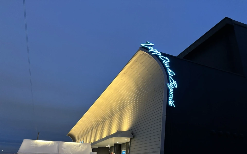 夕暮れの青い空の下、壁が反り返った形のZepp Osaka Bayside。白い壁がライトアップされ、縁に青く光るZepp Osaka Baysideの文字、手前に白いテント