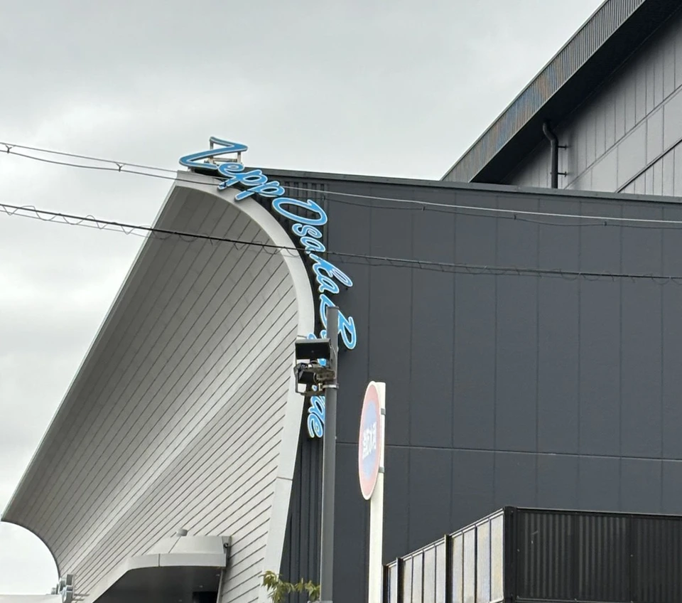 曇り空の下のZepp Osaka Baysideの外観。白い壁が大きく反り返り、その縁に沿って青い筆記体のZepp Osaka Baysideの文字