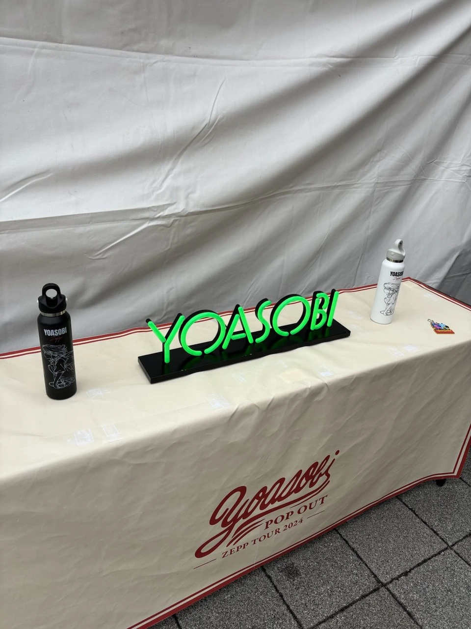 POP OUT ZEPP TOUR 2024と書かれた布をかけた台。黒と白のボトルと、緑に光るYOASOBIのロゴの置物が並んでいる