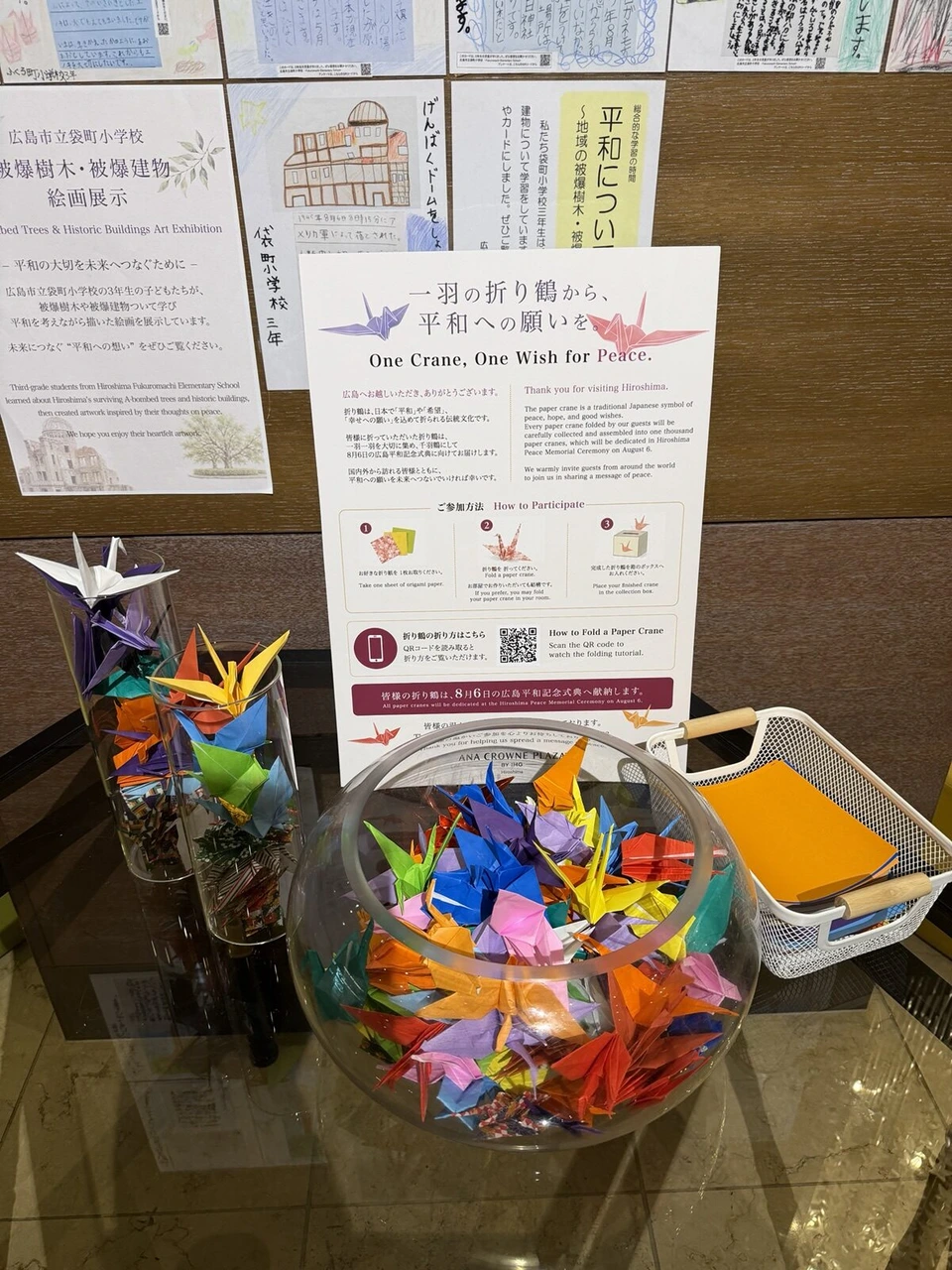 ANAクラウンプラザホテル広島の館内の折り鶴コーナー。ガラスの器に入った折り鶴と案内板、袋町小学校の絵画展示