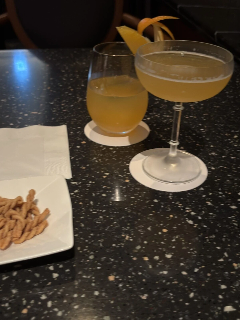 ANAクラウンプラザホテル富山のバーで、黒い石のテーブルに置かれたオレンジ色のカクテル2杯と、ねじったスナック