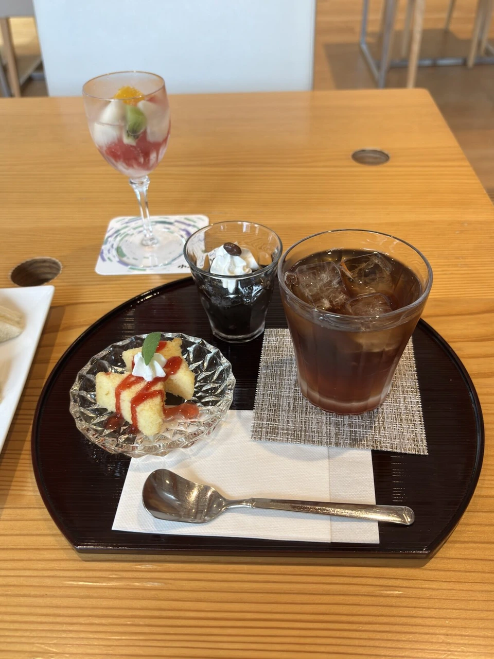 カフェのお盆にのったアイスコーヒー、コーヒーゼリー、いちごソースのカステラ、奥にフルーツのグラス