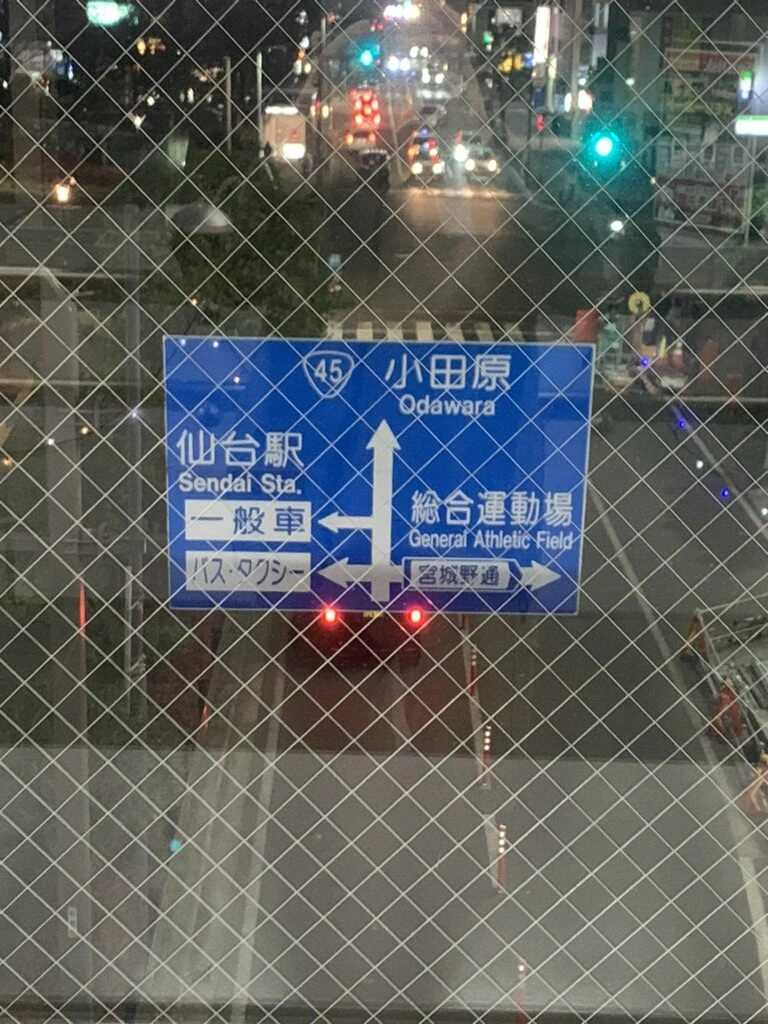 フェンス越しに見た夜の道路と、仙台駅・小田原方面を示す青い案内標識
