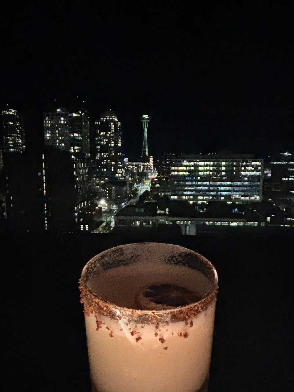 ふちにスパイスをまぶしたグラスのカクテル、後ろに夜景