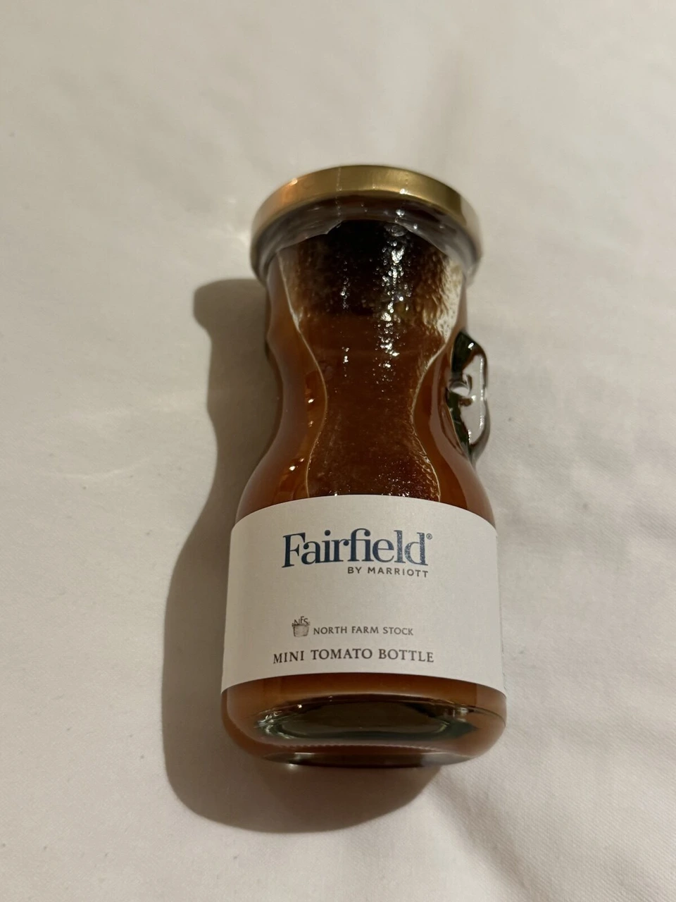 ベッドの上に置いたFairfield BY MARRIOTTのラベルの小瓶MINI TOMATO BOTTLE