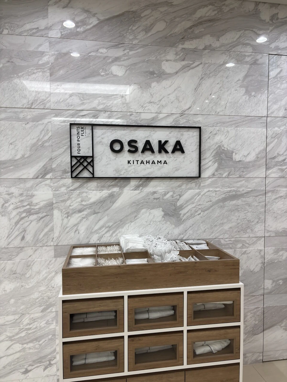 フォーポイント フレックス大阪北浜のロビー。大理石調の壁のOSAKA KITAHAMAのサインと、アメニティの棚