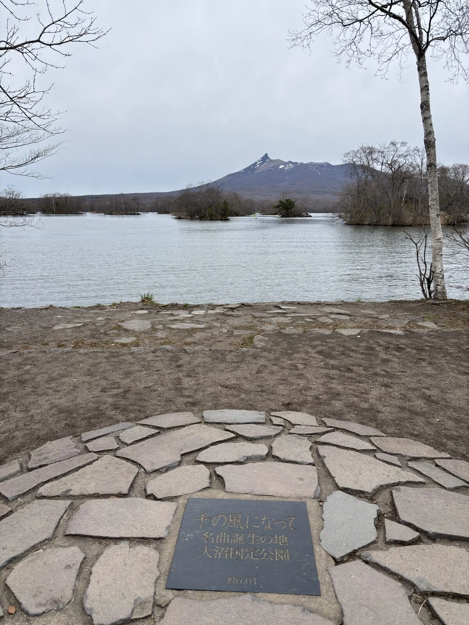 「千の風になって 名曲誕生の地 大沼湖畔公園」のプレートと、湖の向こうの駒ヶ岳