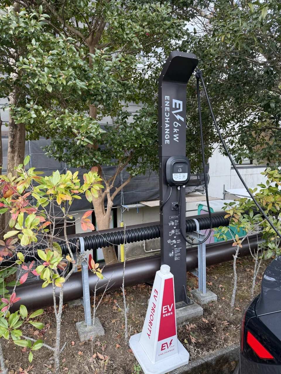 駐車場の植え込みの前に立つEV充電エネチェンジ6kWの充電器と、EV PRIORITY 充電優先のコーン