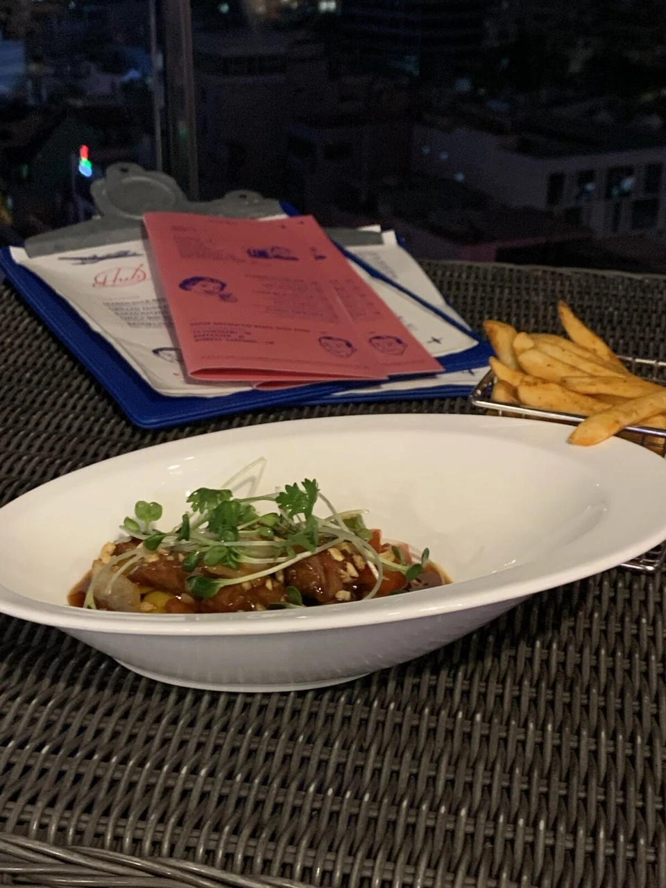 イビス サイゴン エアポートの夜のテラス席のラタンのテーブルに置かれた肉の煮込み料理とフライドポテト、飛行機の絵のメニュー