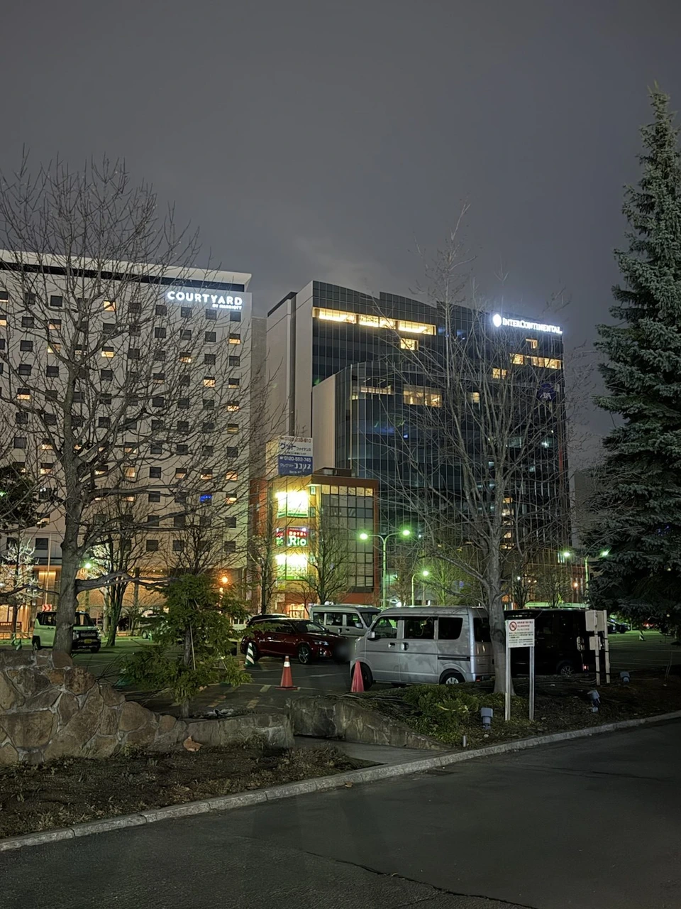 夜のインターコンチネンタル札幌のまわり。右に最上部でINTERCONTINENTALの文字が光るホテル、左にCOURTYARDの看板のビル、手前に駐車場と冬枯れの木