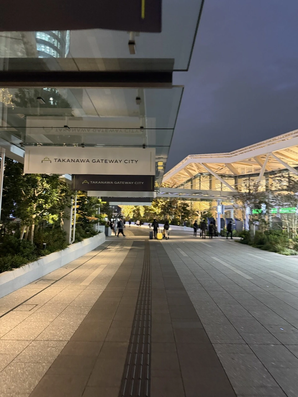 TAKANAWA GATEWAY CITYのサインが下がる歩道と、奥の高輪ゲートウェイ駅の屋根