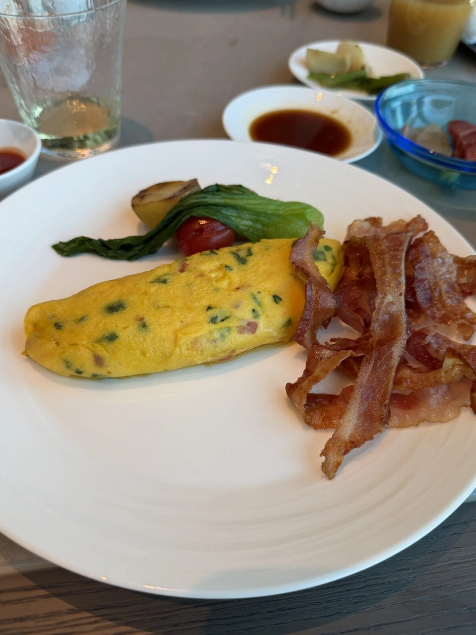 ほうれん草とハムのオムレツにカリカリのベーコン、焼いた小玉ねぎ、トマト、青菜。奥に小鉢と醤油の皿