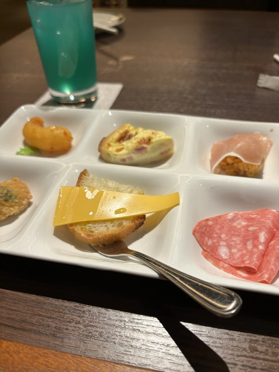 仕切られた皿にチーズ、サラミ、生ハム、キッシュと青いドリンク