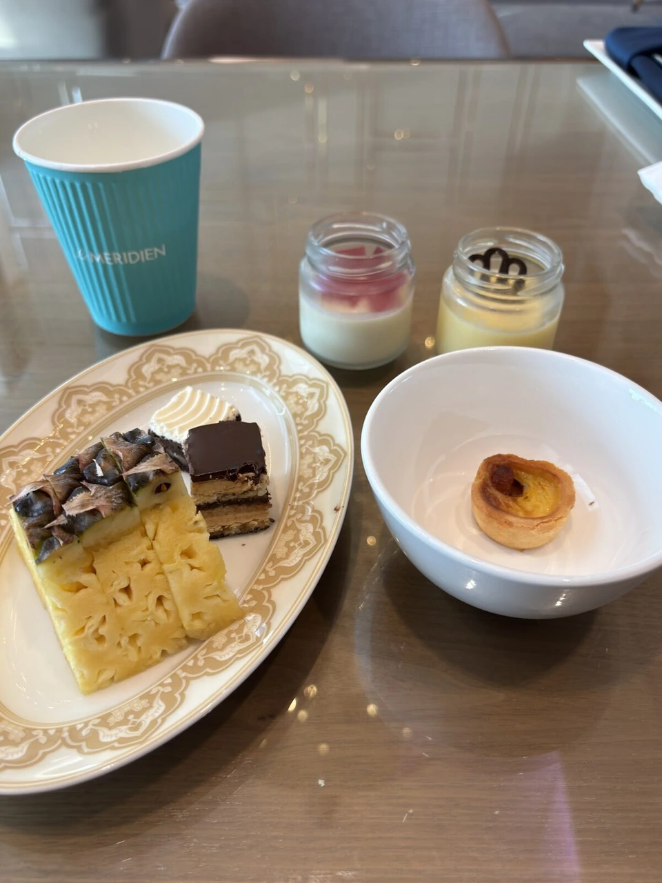 ラウンジのデザート。パイナップルケーキのようなケーキ、チョコレートケーキ、プリン2つ、エッグタルトと、Le MERIDIENのロゴ入りの水色の紙コップ