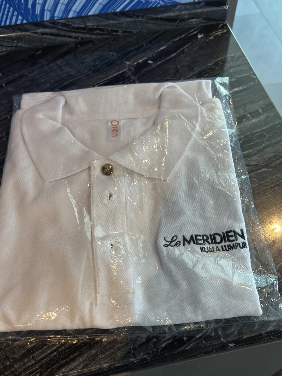 黒い大理石のテーブルの上の、袋に入った白いポロシャツ。胸にLe MERIDIEN KUALA LUMPURのロゴ