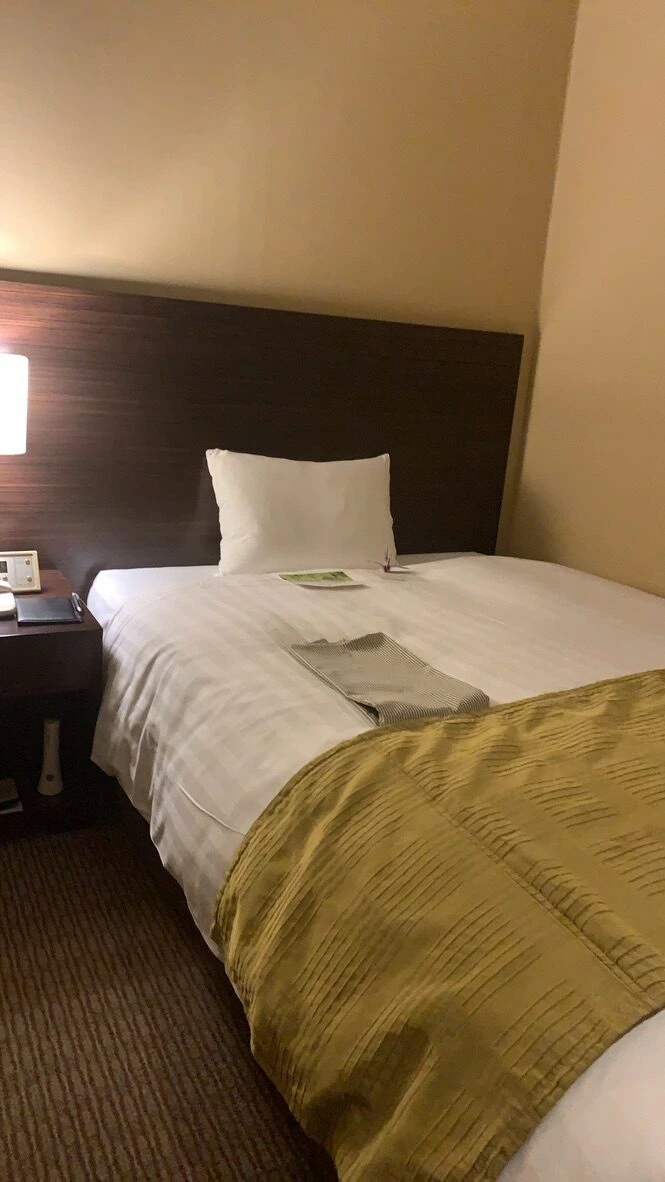 ホテル日航つくばのシングルルーム。白いシーツのベッドにオリーブ色のベッドスロー、木目のヘッドボードと読書灯