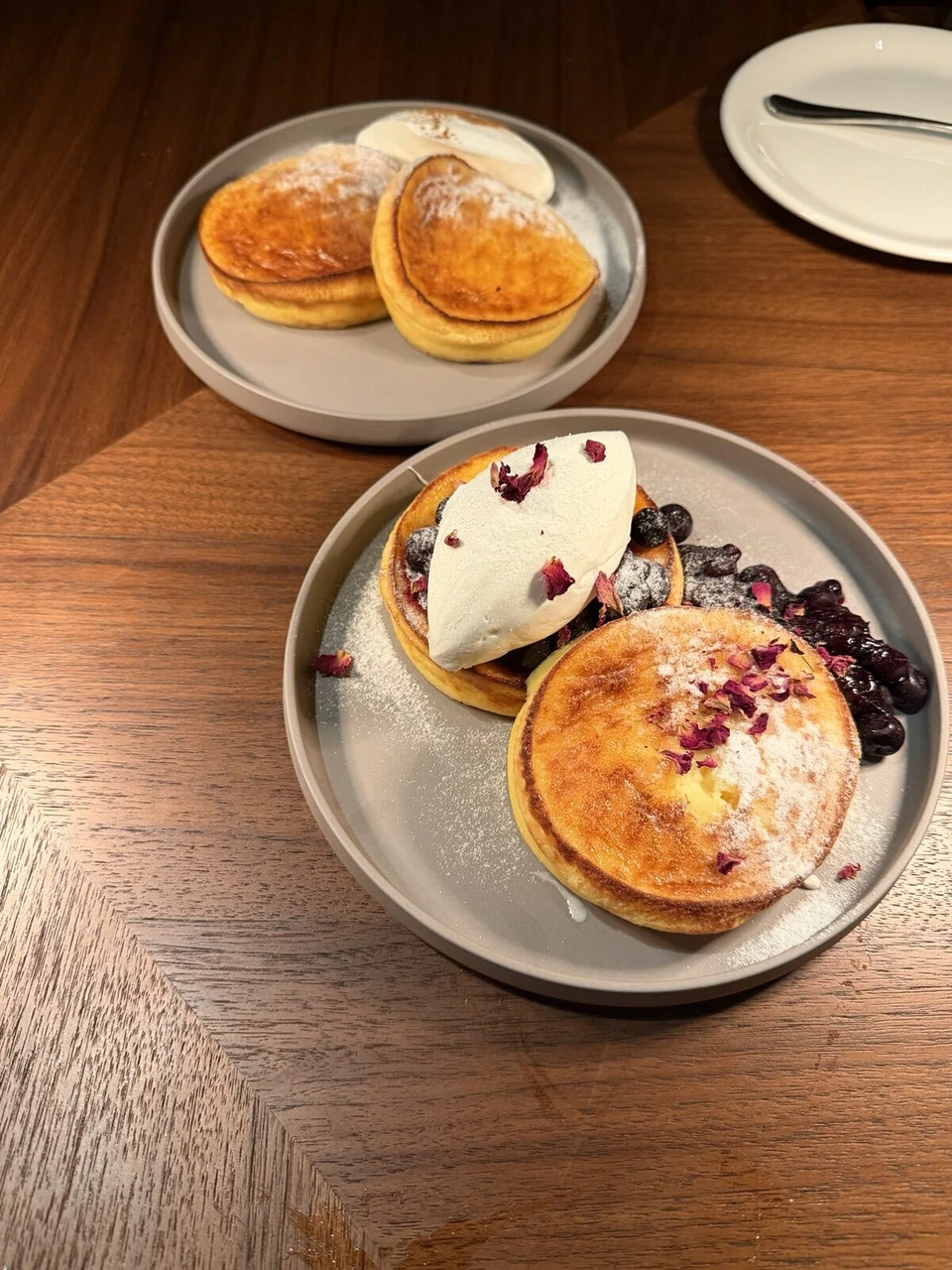 木のテーブルに置かれたパンケーキ2皿。片方にクリームとベリー、バラの花びら