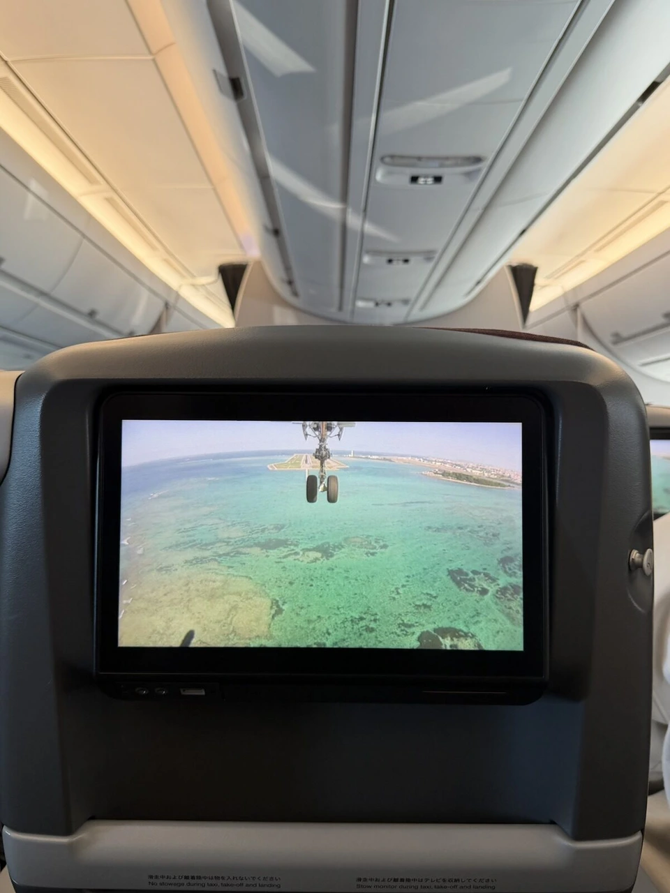 座席モニターに映る着陸カメラの映像。脚の下にエメラルド色のサンゴ礁と那覇空港の滑走路