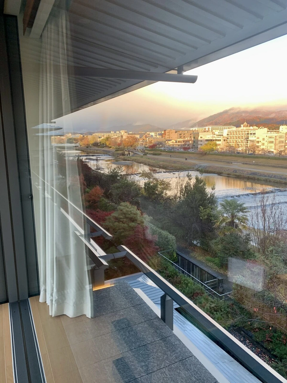 カーテン越しに見たバルコニーと鴨川。夕日で対岸の建物と山が明るい