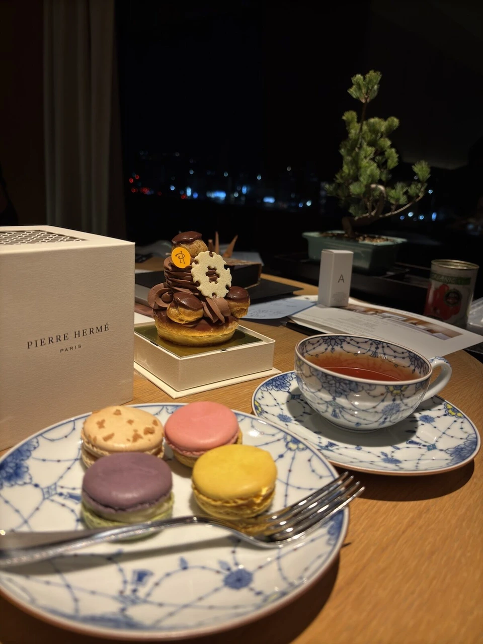 窓際のテーブルのマカロン、チョコのケーキ、紅茶と盆栽。窓の外は夜景