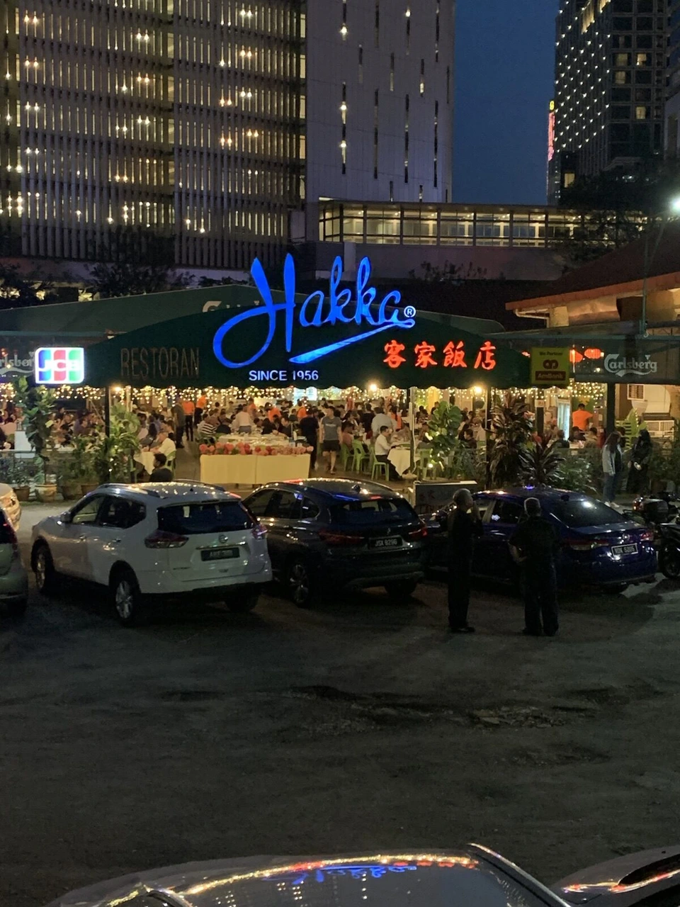 夜の客家飯店。青いHakkaのネオンと電飾、店の前に並ぶ車