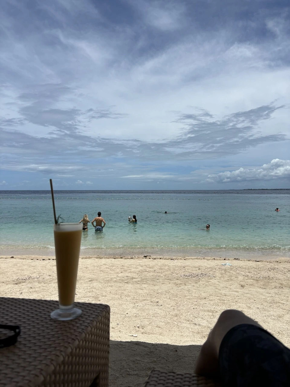 ビーチのサイドテーブルに置いたジュースと、遠浅の海で泳ぐ人