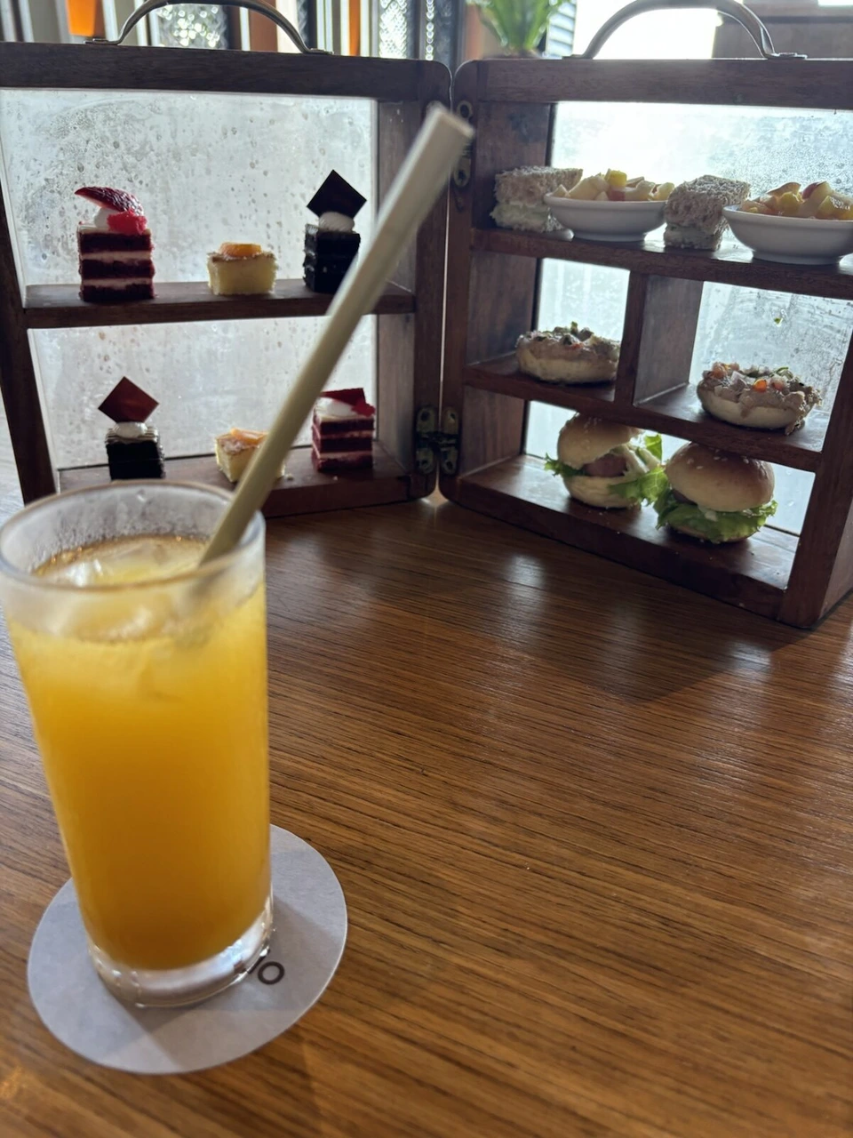 ストロー付きのオレンジジュースと、奥に置かれた木の箱に並ぶケーキとミニバーガー