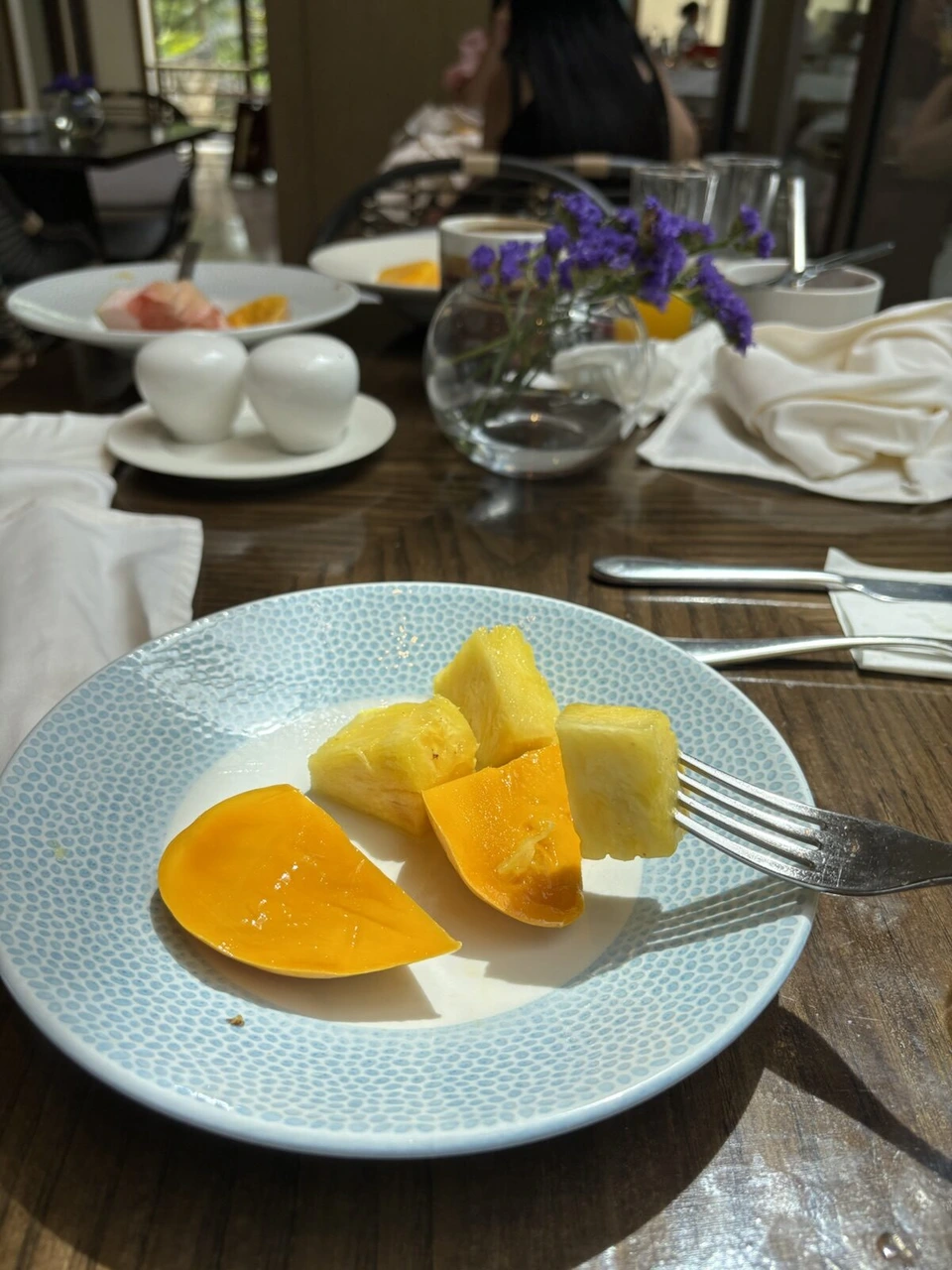 朝食会場のテーブル。水色の皿にのったマンゴーとパイナップル、奥に紫の花