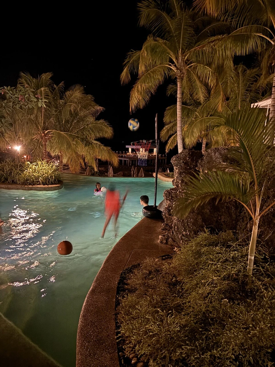 夜のプール。ヤシの木の間でボール遊びをする人